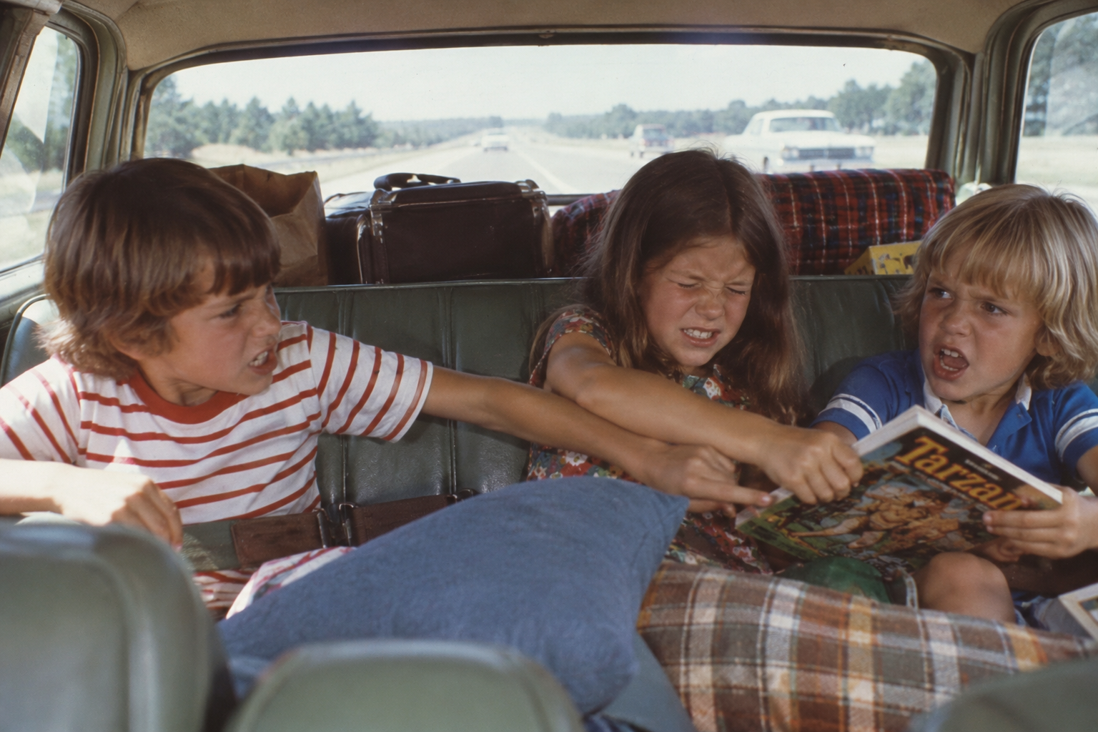 Three children argue and pull at a Tarzan comic book in the backseat of a packed car during a road trip, with suitcases and a blanket nearby.