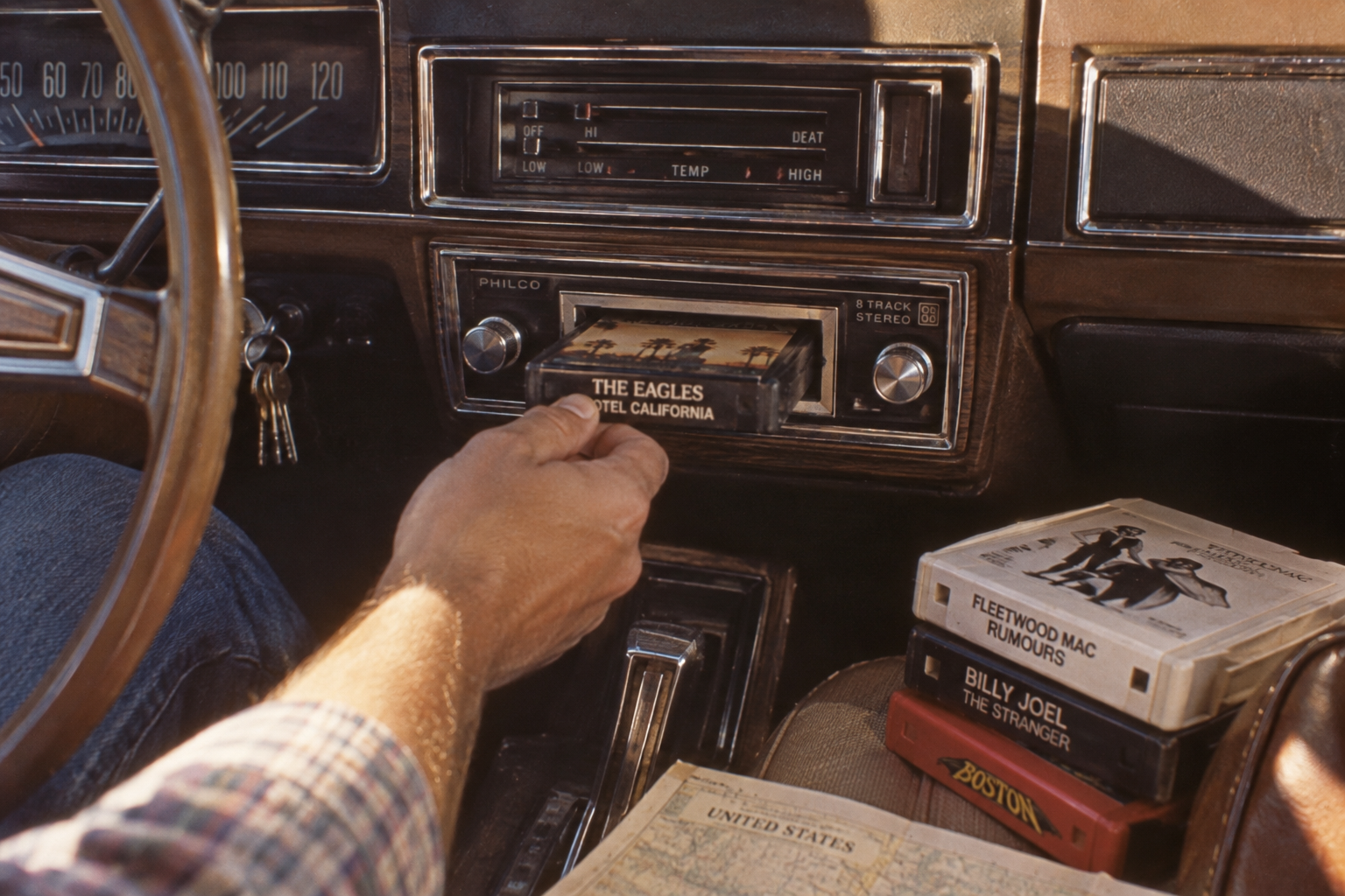 A person inserts an Eagles “Hotel California” 8-track tape into a car stereo. Other 8-tracks—Fleetwood Mac, Billy Joel, and Boston—plus a map, rest nearby. The car dashboard and steering wheel are visible.