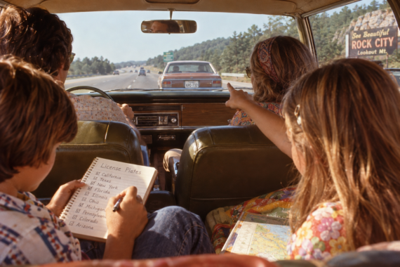 A family drives down a highway, kids in the backseat play a license plate game. One child writes on a clipboard, another points ahead, and a Rock City billboard is visible outside.