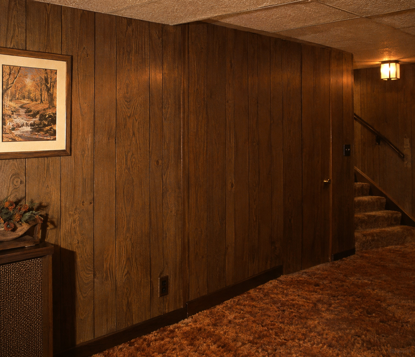 A dimly lit room with dark wood-paneled walls, a brown carpeted floor, a framed landscape painting, stairs leading up, and a small table with a plant arrangement. 1970s basement.