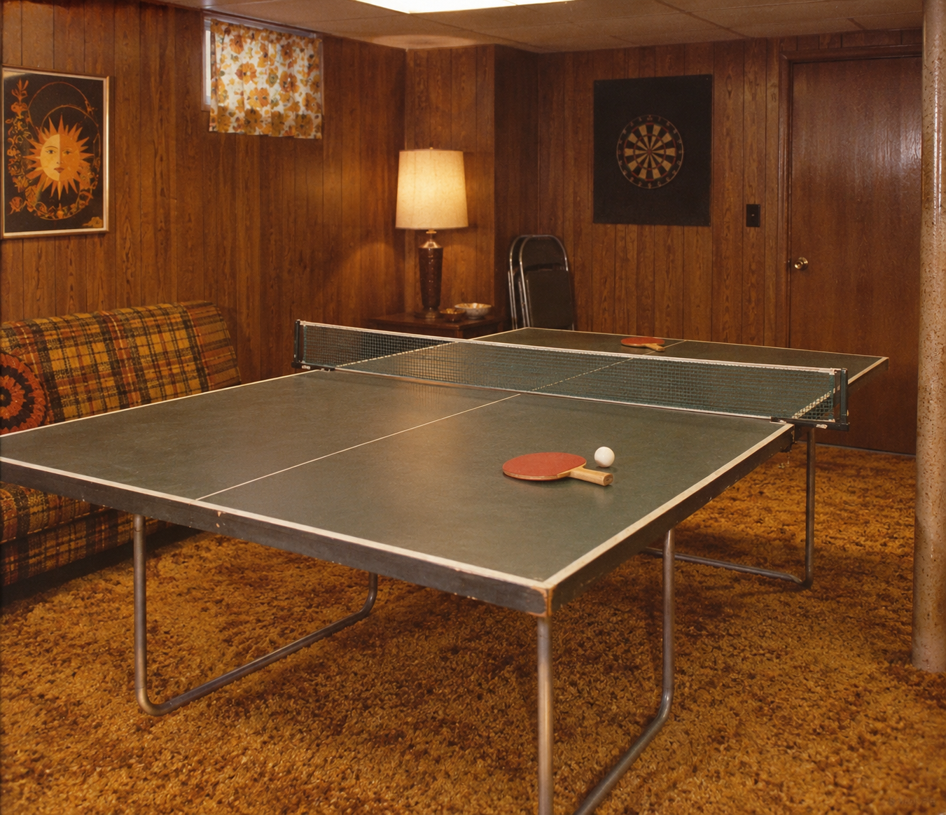 A vintage basement with wood-paneled walls, a plaid couch, a lamp, dartboard, and a ping pong table set up with paddles and a ball. Carpet is brown and decor is retro-style.