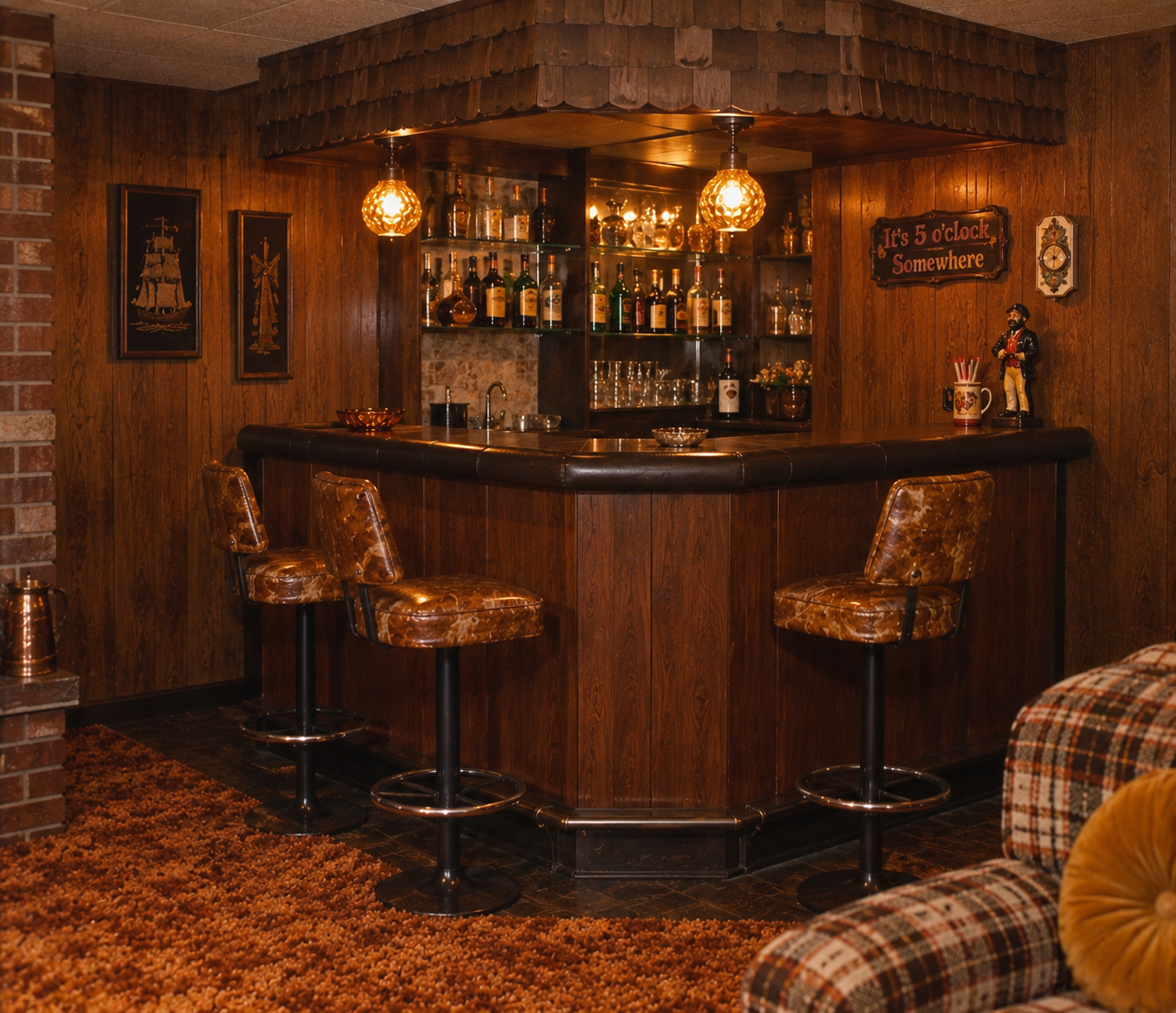 Retro basement bar with wood paneling, three brown barstools, glass shelves of liquor, hanging lights, and a sign reading “It’s 5 o’clock Somewhere.” Plaid couch and orange carpet are also visible.