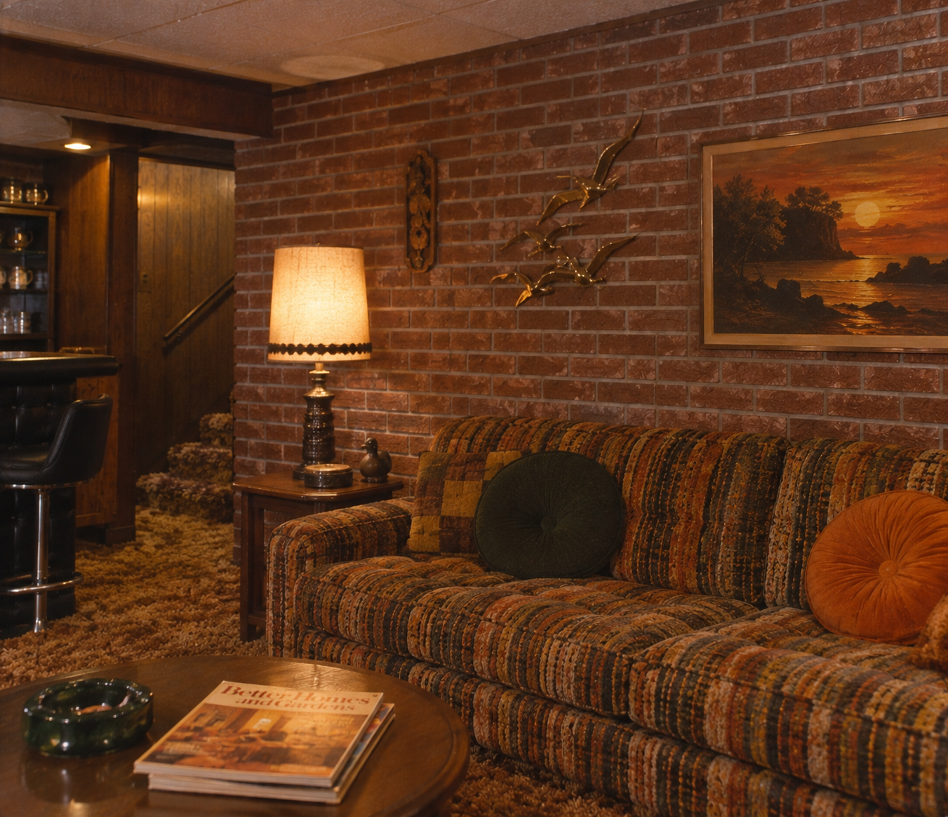 A retro living room with a patterned sofa, round pillows, a brick wall, a glowing lamp, sunset artwork, gold bird décor, and a round table with magazines and a green ashtray. Stairs and a bar area are in the background.