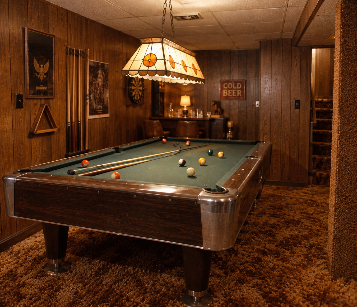 A vintage basement with wood-paneled walls, a pool table with balls and cues, a stained-glass light, dartboard, posters, and a “COLD BEER” sign. Carpeted floor and stairs are visible in the background.