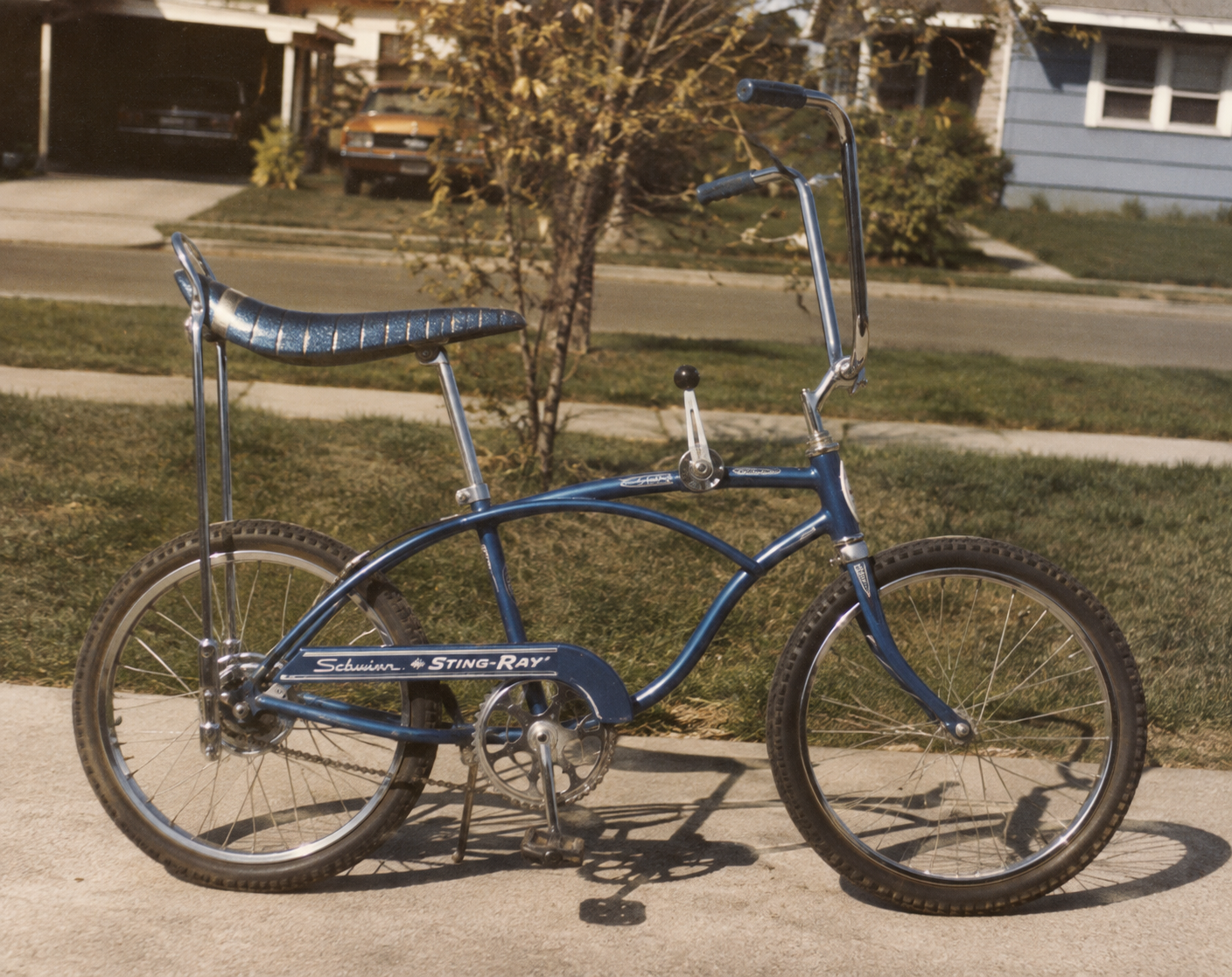 A vintage blue Schwinn Sting-Ray bicycle with a banana seat and high-rise handlebars is parked on a driveway in front of a suburban house and lawn.