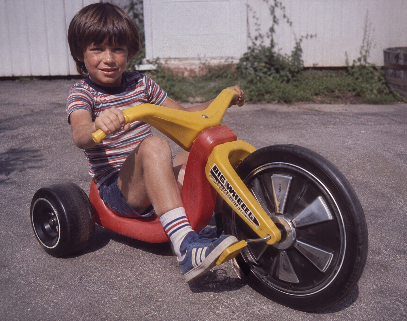 A young boy in striped shirt and shorts rides a large plastic tricycle with a yellow frame and oversized front wheel on a paved driveway.