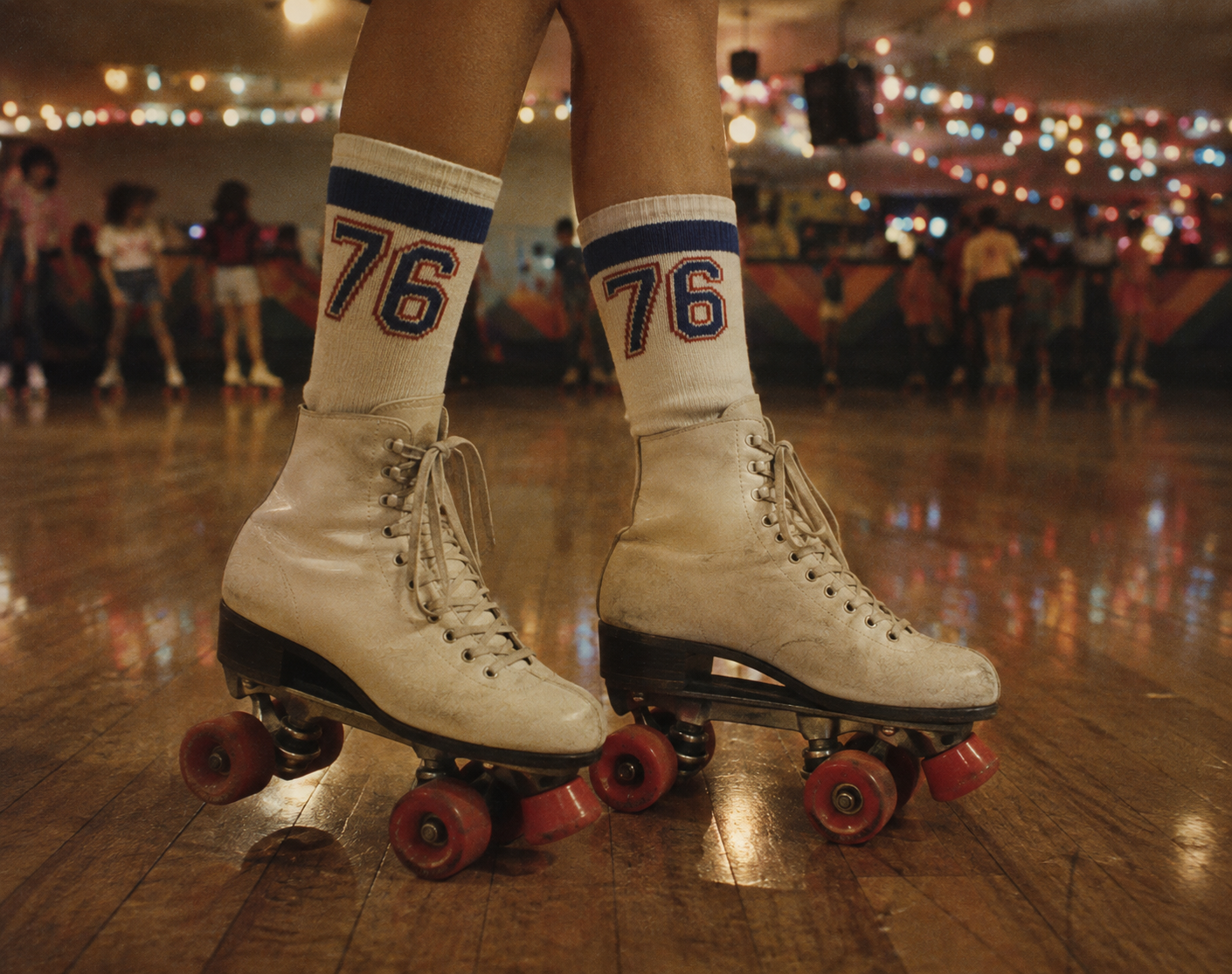 Close-up of a person wearing vintage white roller skates and socks with “76” on them, standing on a shiny wooden rink floor with colorful lights and skaters blurred in the background.
