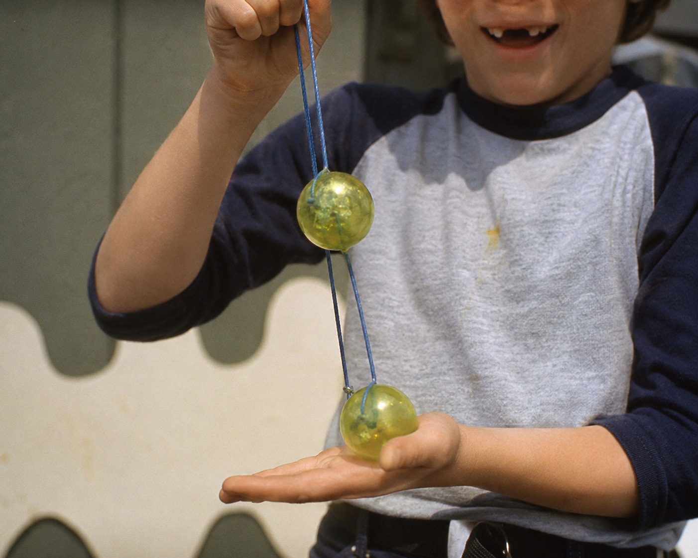 A child in a baseball-style shirt plays with two yellow-green balls on strings, holding one above the other while smiling.