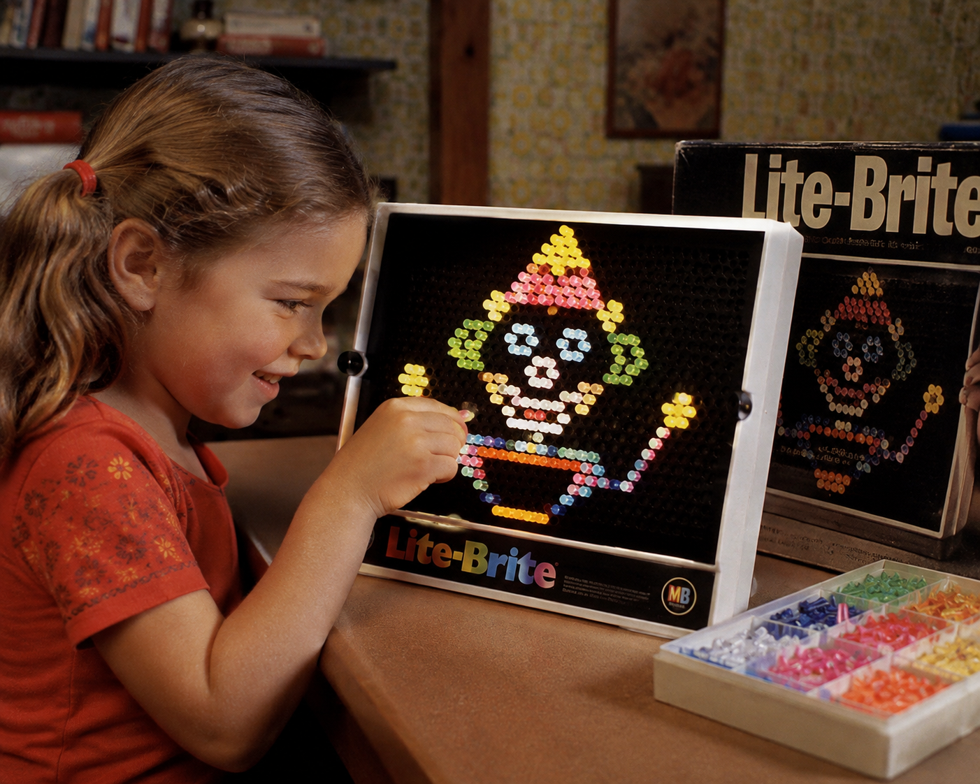 A young girl smiles as she places colorful pegs into a Lite-Brite, creating a clown design. Lite-Brite box and peg trays are beside her on the table in a cozy room.
