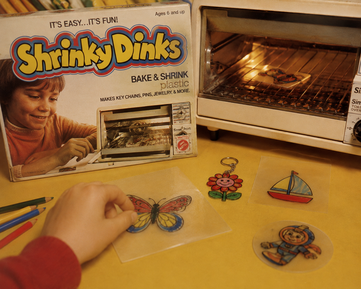 A hand holds decorated Shrinky Dinks plastic, with a butterfly design near a toaster oven. Finished Shrinky Dinks—a flower, boat, and bear—lie on a yellow surface. The Shrinky Dinks box is visible in the background.