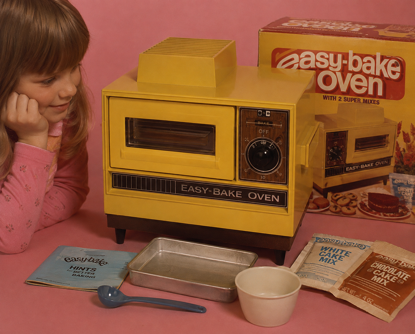 A young girl smiles at a yellow Easy-Bake Oven with mixes, a pan, spoon, bowl, and the box displayed on a pink background.