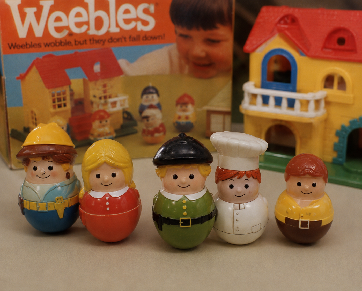 Five colorful Weebles toys dressed as various characters stand in a row. A toy house and the original Weebles box, showing a smiling child, are visible in the background.