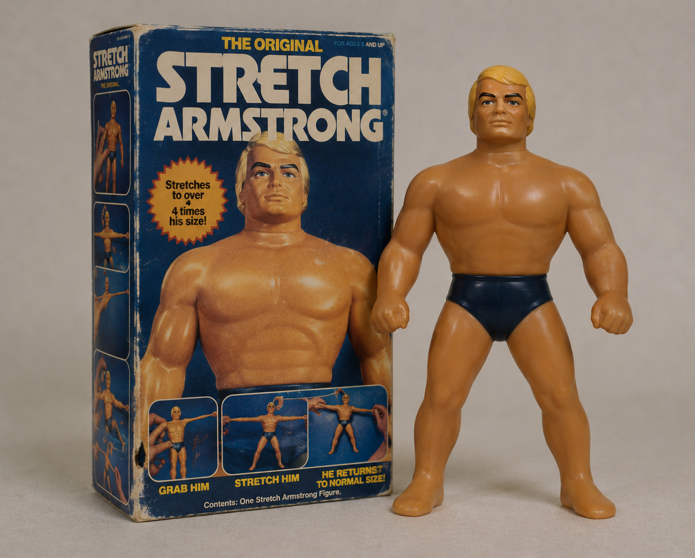 Stretch Armstrong action figure standing next to its original box, which features images and details about the toy’s stretching abilities. The figure wears black trunks and has blonde hair.