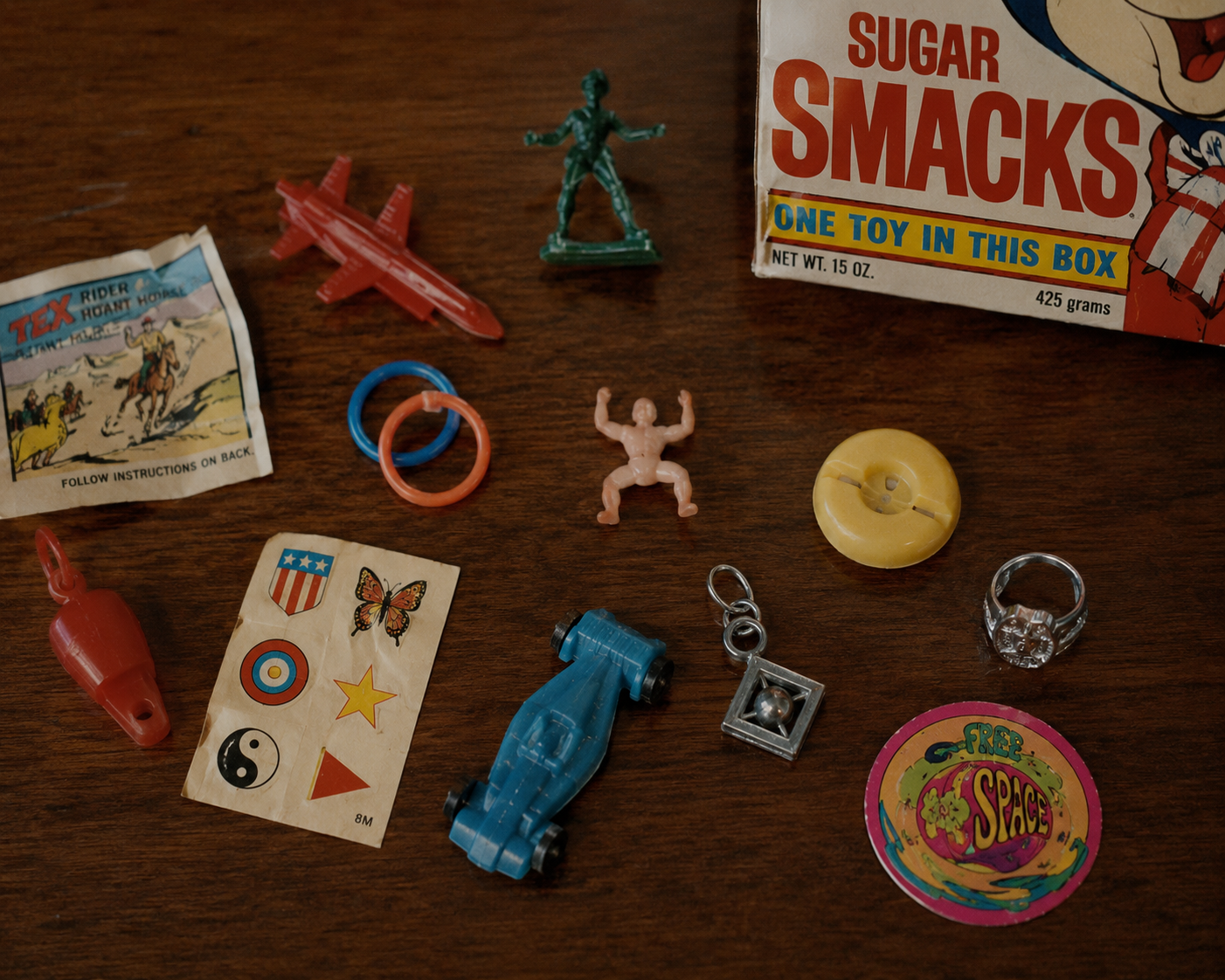 A collection of vintage cereal box toys, including a toy car, whistle, plastic rings, figurines, charms, stickers, and a Sugar Smacks cereal box, displayed on a wooden surface.