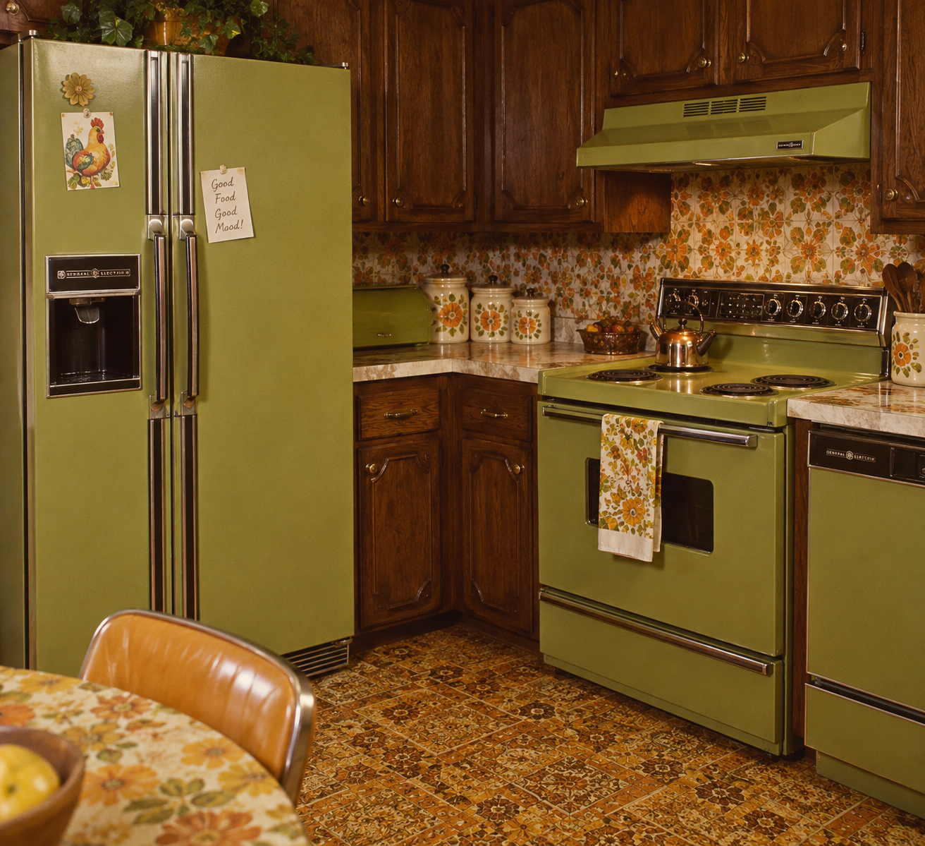 A vintage kitchen with olive green appliances, floral patterned wallpaper, and brown wooden cabinets. The fridge has notes, and floral tins line the counter. An orange floral towel hangs from the stove. 1970s home features.