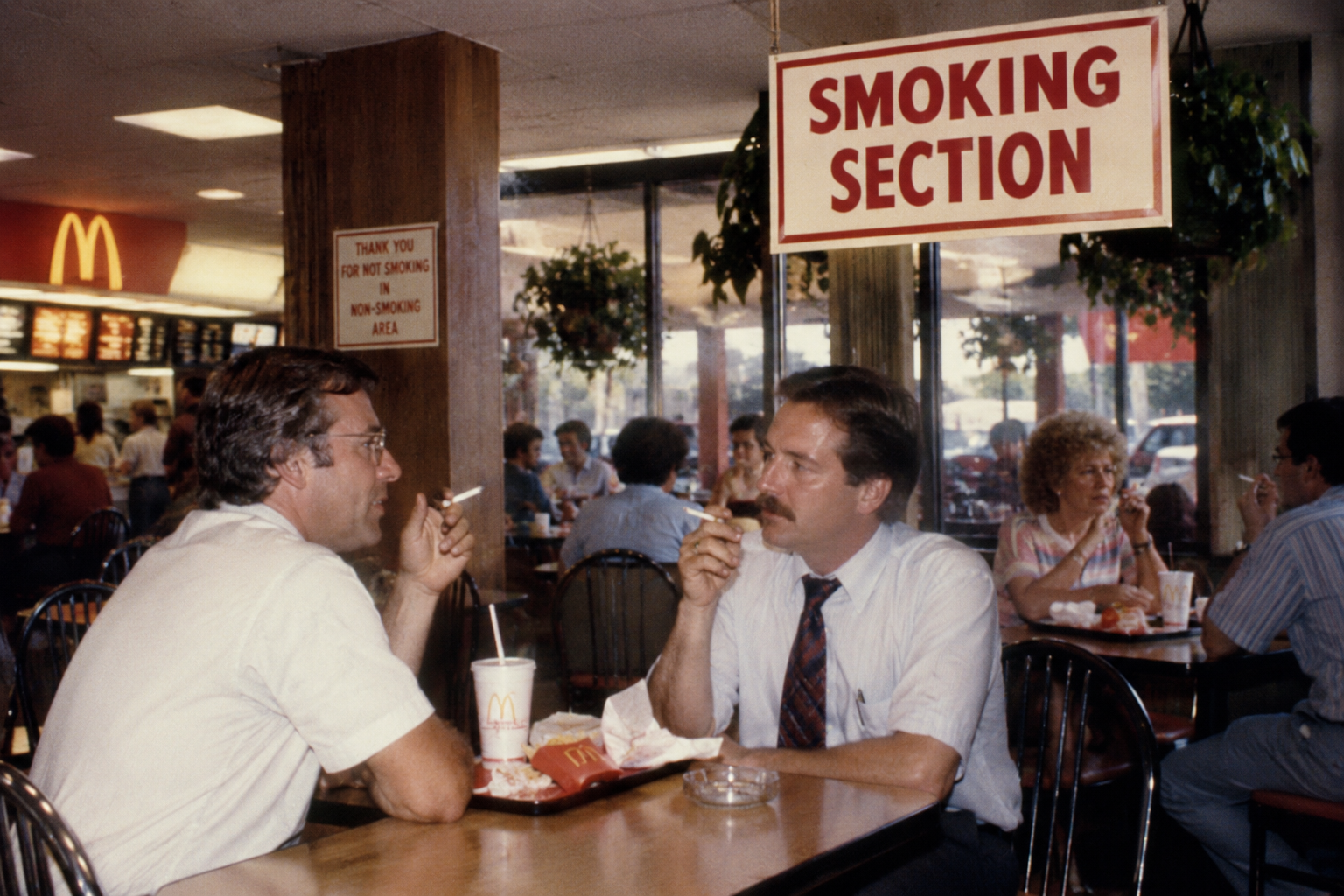 Two men in business attire sit at a McDonald’s table under a “Smoking Section” sign, smoking cigarettes. Other customers are eating in the busy restaurant. A McDonald’s meal with fries and a drink is on the table. ’80s fast-food restaurant features.