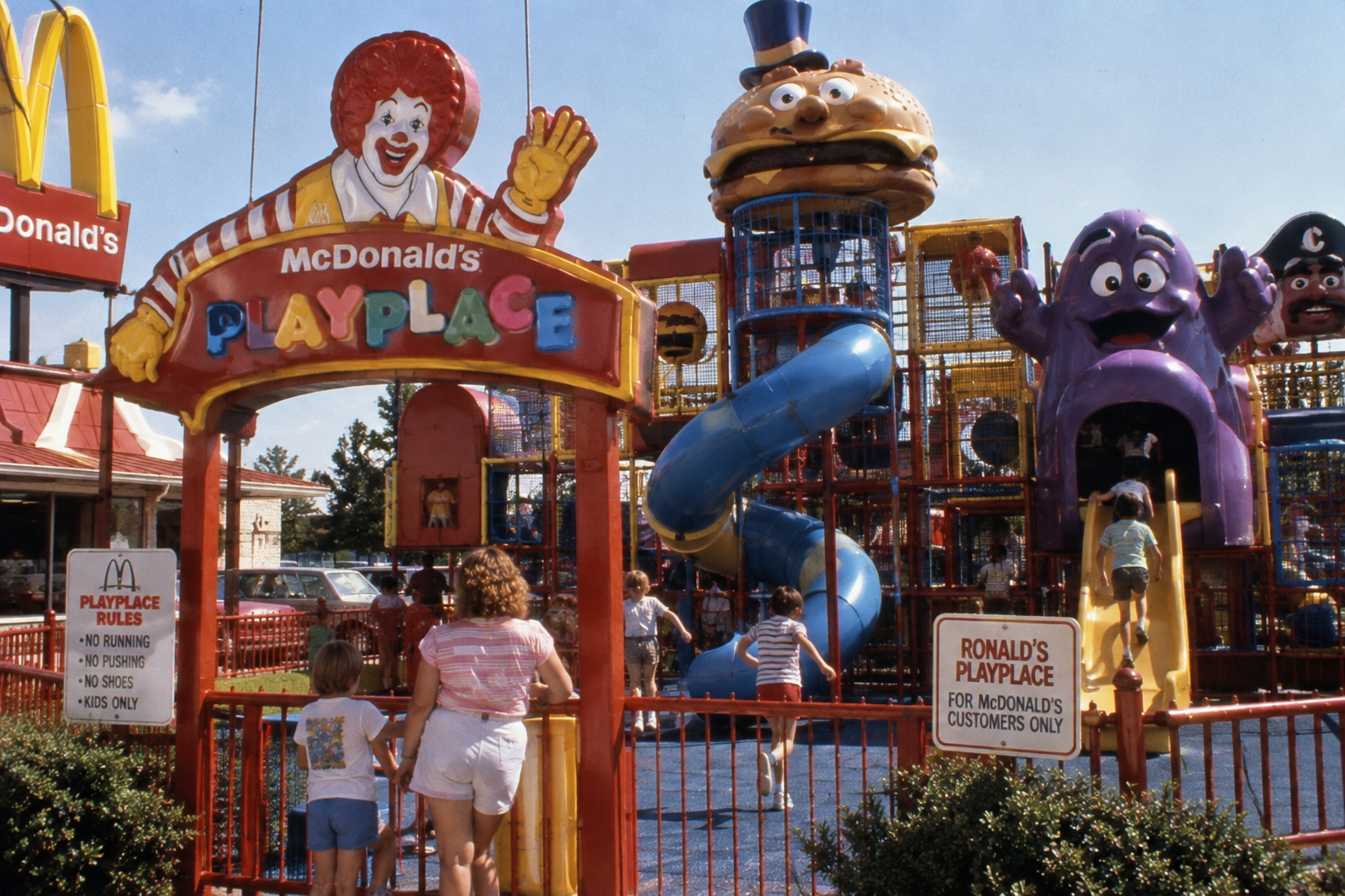 Children play at a colorful McDonald’s PlayPlace with slides and climbing structures shaped like characters and food items, under a sunny sky. Signs display playground rules.