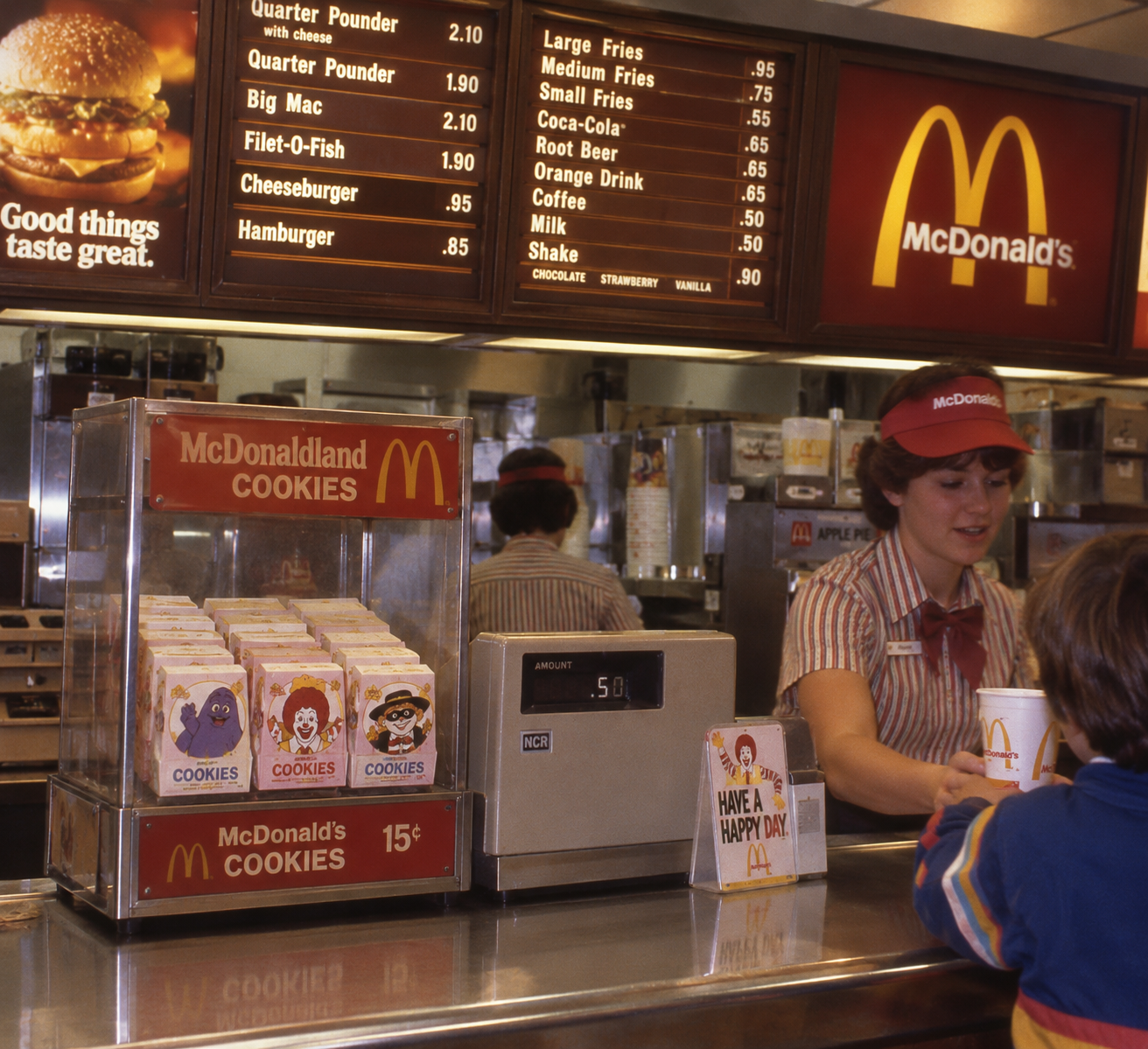 A McDonald’s employee serves a child at the counter. Behind her, a menu board lists food prices. Packs of McDonaldland Cookies are displayed next to a cash register showing $5.00.