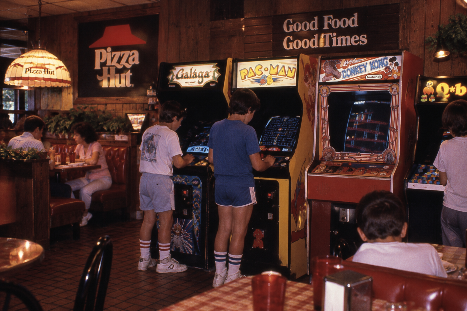 Kids play Galaga, Pac-Man, and Donkey Kong arcade games inside a vintage Pizza Hut restaurant, with booths and people dining in the background. A sign reads “Good Food, Good Times.”