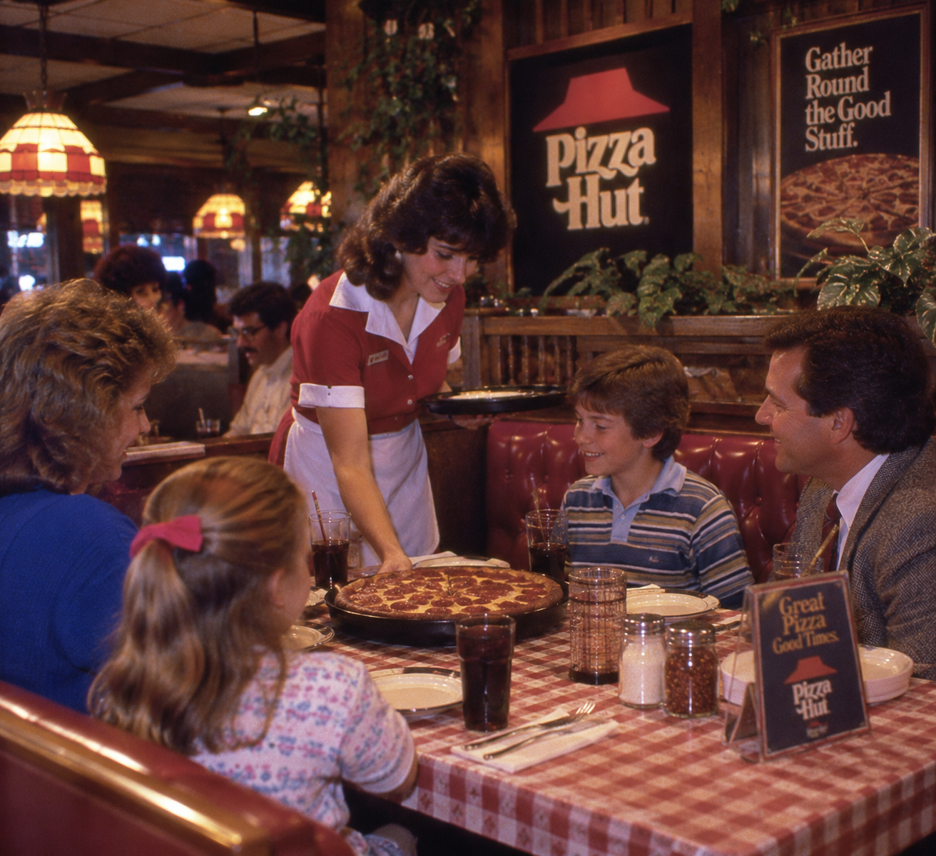 A waitress serves pizza to a smiling family of four at a Pizza Hut restaurant, with checkered tablecloths and classic 1980s decor.