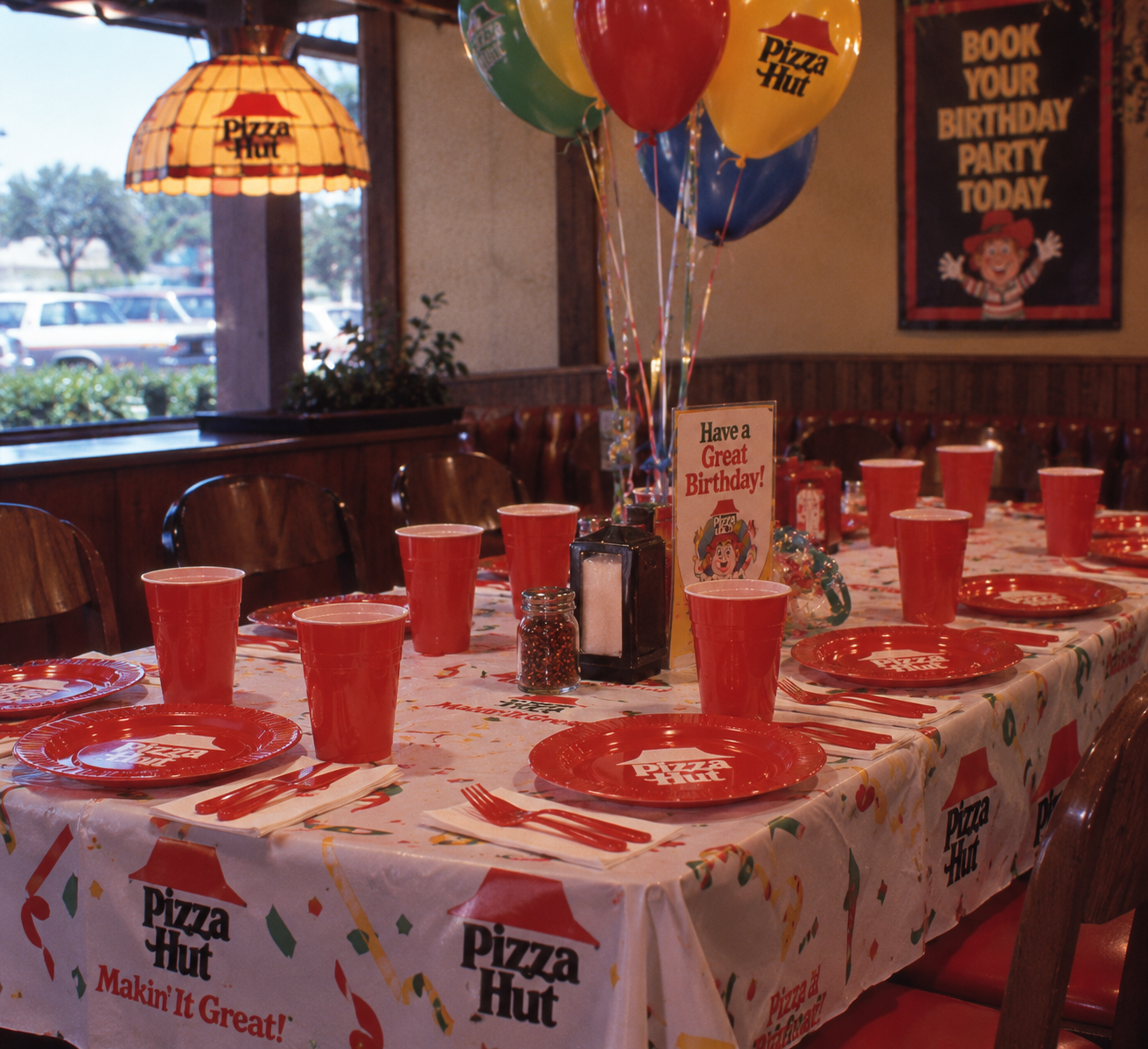 A Pizza Hut birthday party table is set with red plates, cups, colorful balloons, confetti tablecloth, and birthday decorations. A sign says “Have a Great Birthday!” with a party menu and a poster in the background.
