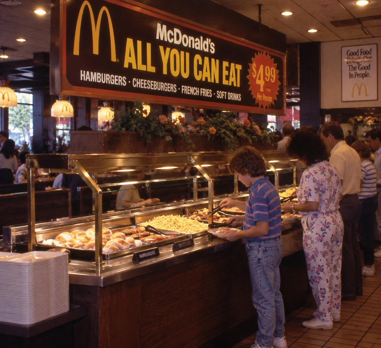People serve themselves food at a McDonald’s all-you-can-eat buffet. There is a large yellow sign advertising the buffet, and several trays of different foods are visible on the counter.