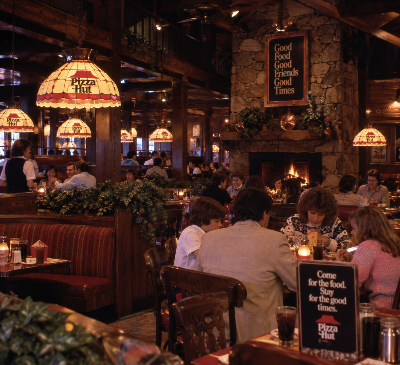 A cozy, dimly lit Pizza Hut restaurant filled with diners, wooden booths, stained glass lamps, a stone fireplace, and signs reading “Good Food, Good Friends, Good Times.”