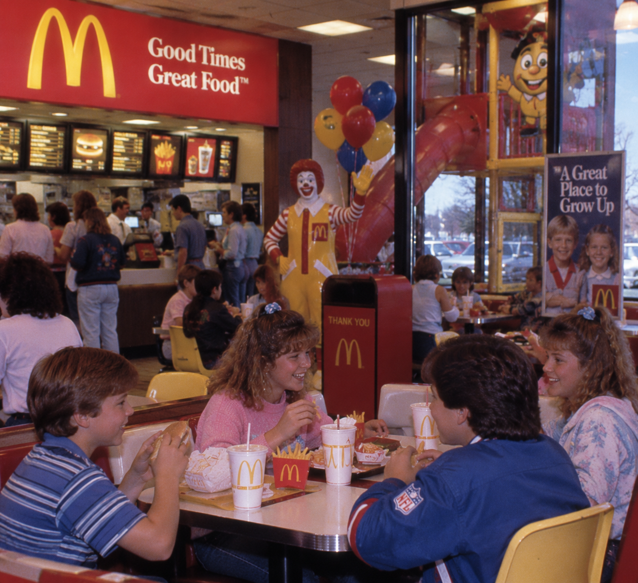 Four kids eat McDonald’s food at a table inside a busy restaurant. The counter and Ronald McDonald statue are visible in the background along with a sign reading “Good Times Great Food.”