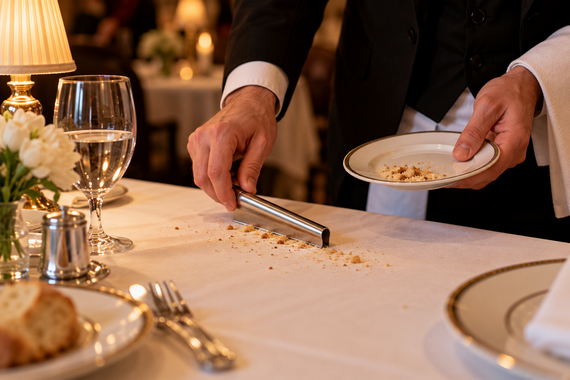 A server in formal attire uses a metal scraper to clear bread crumbs from a white tablecloth in an elegant, candlelit restaurant.