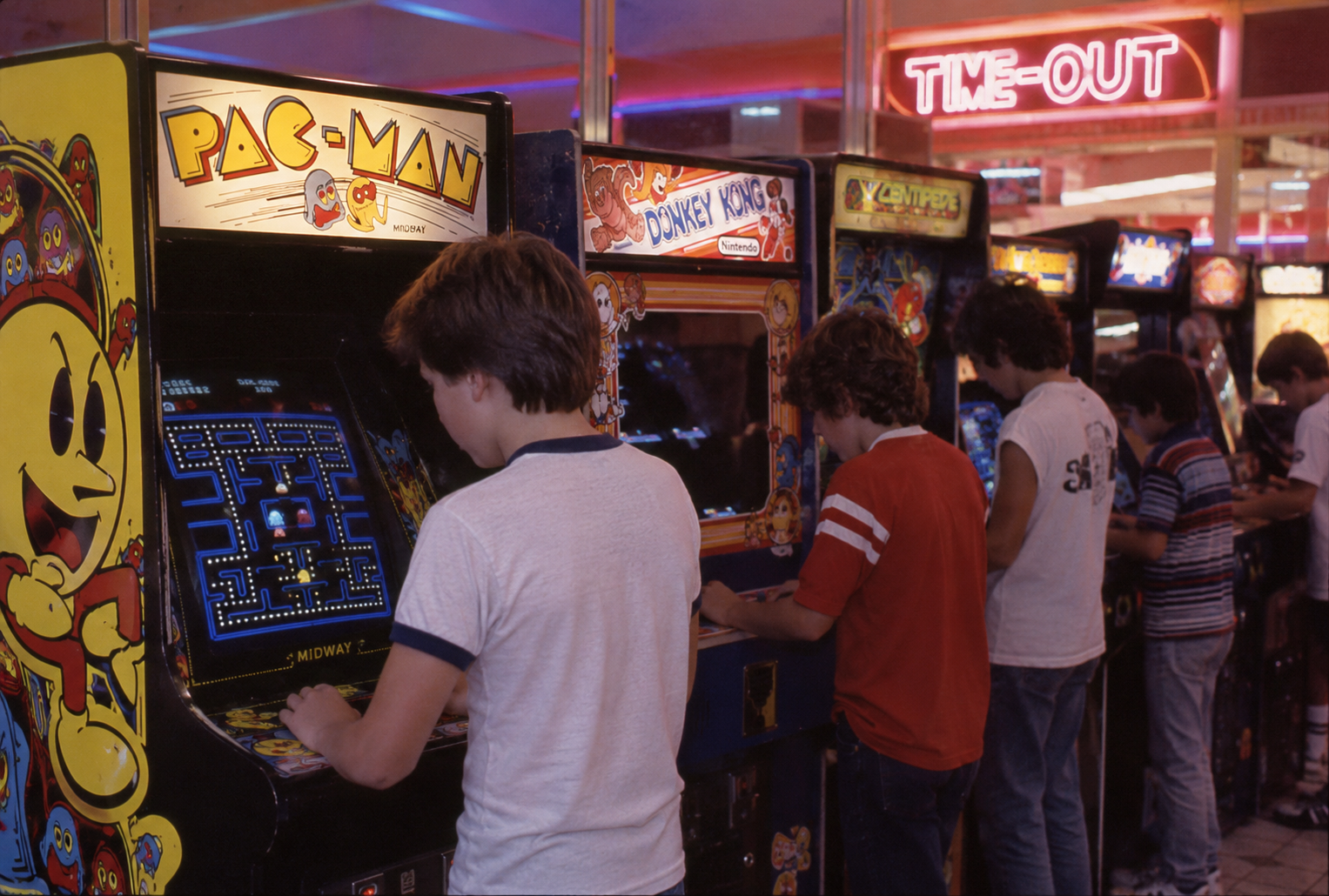 Teenagers play classic arcade games like Pac-Man, Donkey Kong, and Centipede in a retro arcade with neon lights and a “TIME-OUT” sign glowing in the background. ’80s mall activities.