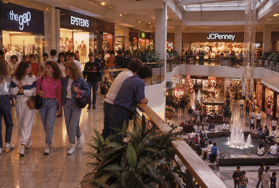 Shoppers walk through a busy 1980s mall with stores like the Gap, Epsteins, and JCPenney. People gather near a fountain and plants, and the atmosphere is lively and crowded.