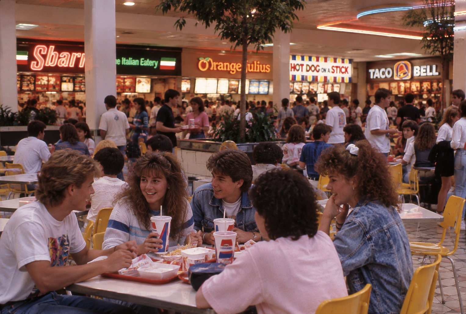 A group of teens sit at a table in a busy food court, eating and laughing. Behind them are fast food counters, including Sbarro, Orange Julius, Hot Dog on a Stick, and Taco Bell.