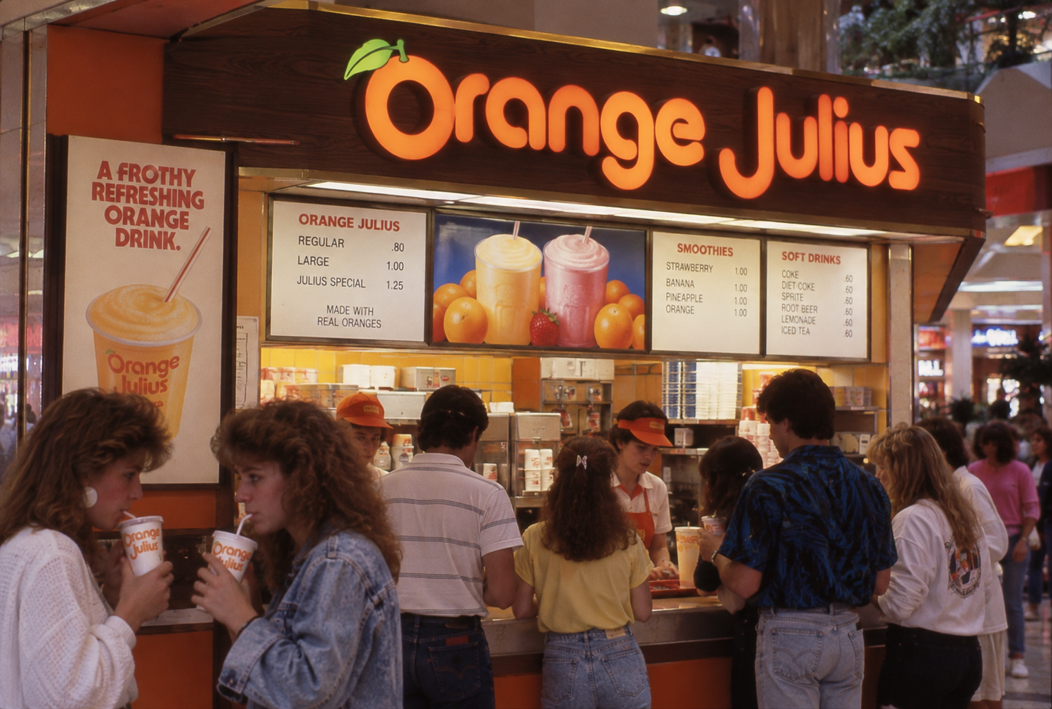 People stand at an Orange Julius counter in a mall, ordering and drinking beverages. The counter is bright, with a large menu displaying drink options like smoothies and soft drinks. The atmosphere is busy and lively.