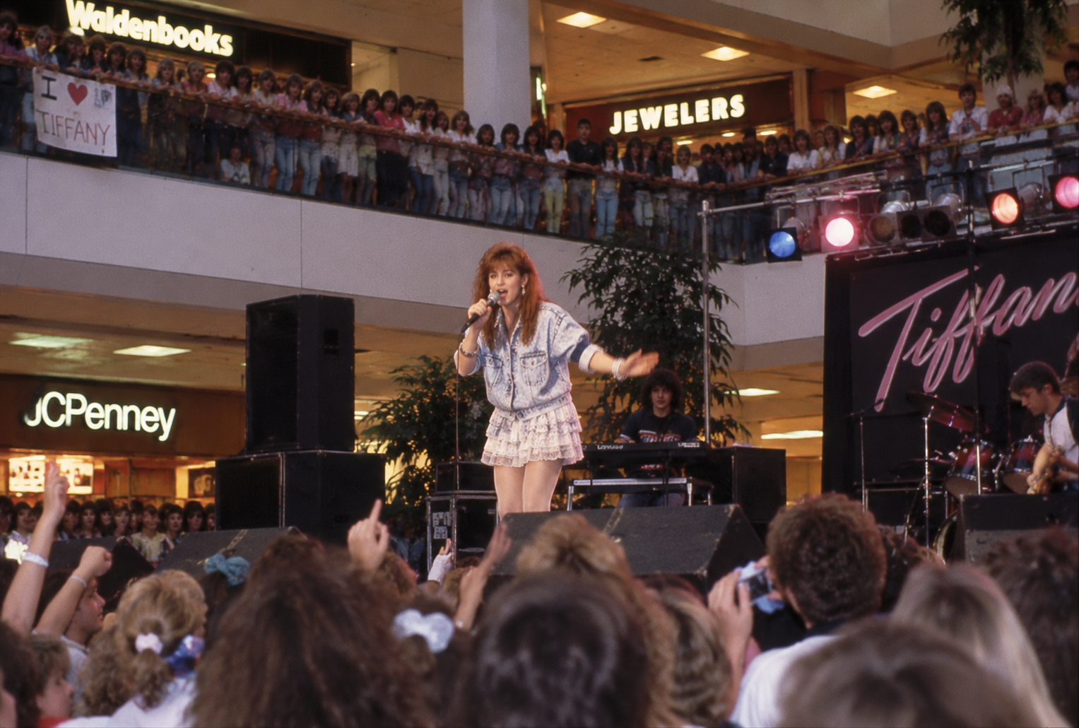 A singer performs on stage at a busy shopping mall, with crowds cheering and a banner reading “Tiffany” behind her. Store signs for Waldenbooks, Jewelers, and JCPenney are visible in the background.