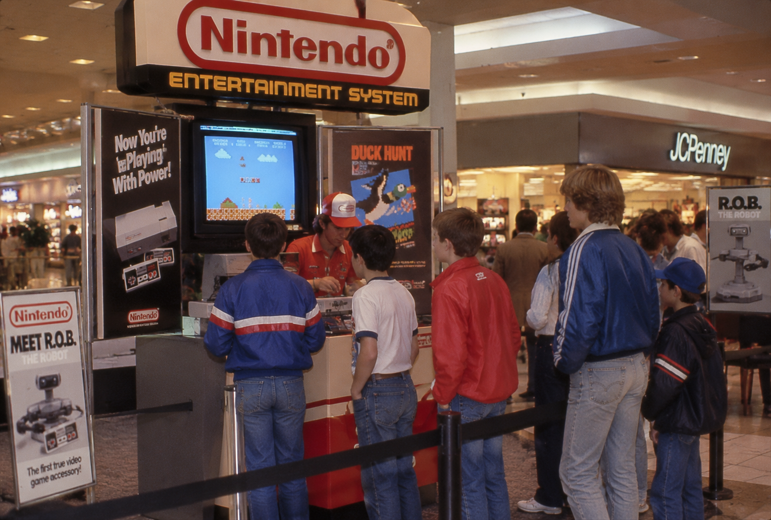 A group of kids watches a Nintendo Entertainment System demo at a mall display. A person in a red hat explains as Super Mario Bros. plays on the screen. JC Penney is visible in the background.