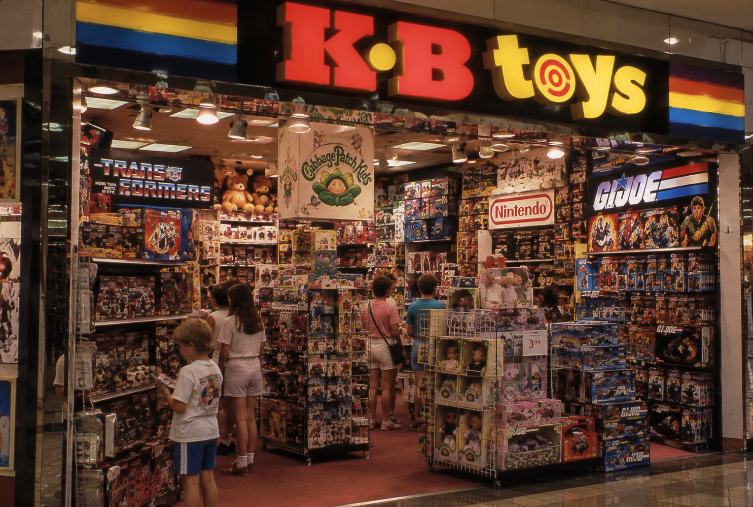 Children and adults browse toys in a brightly lit KB Toys store filled with action figures, plush toys, and games. The store signage is colorful and shelves are packed with recognizable brands like Transformers and Nintendo.