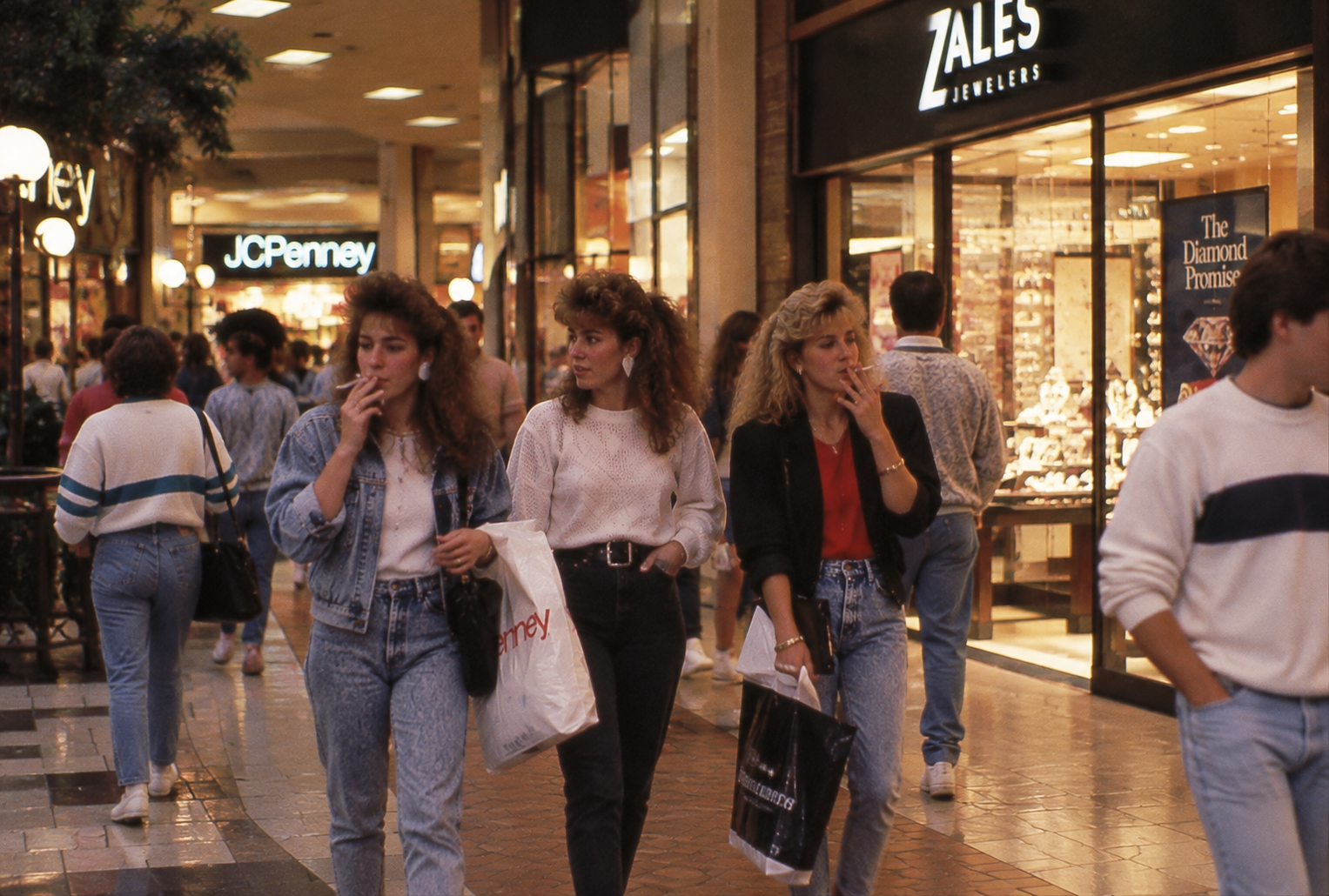 Three women in 1980s clothing walk through a shopping mall, carrying bags and smoking. Other shoppers pass by, and store signs for JC Penney and Zales Jewelers are visible in the background.