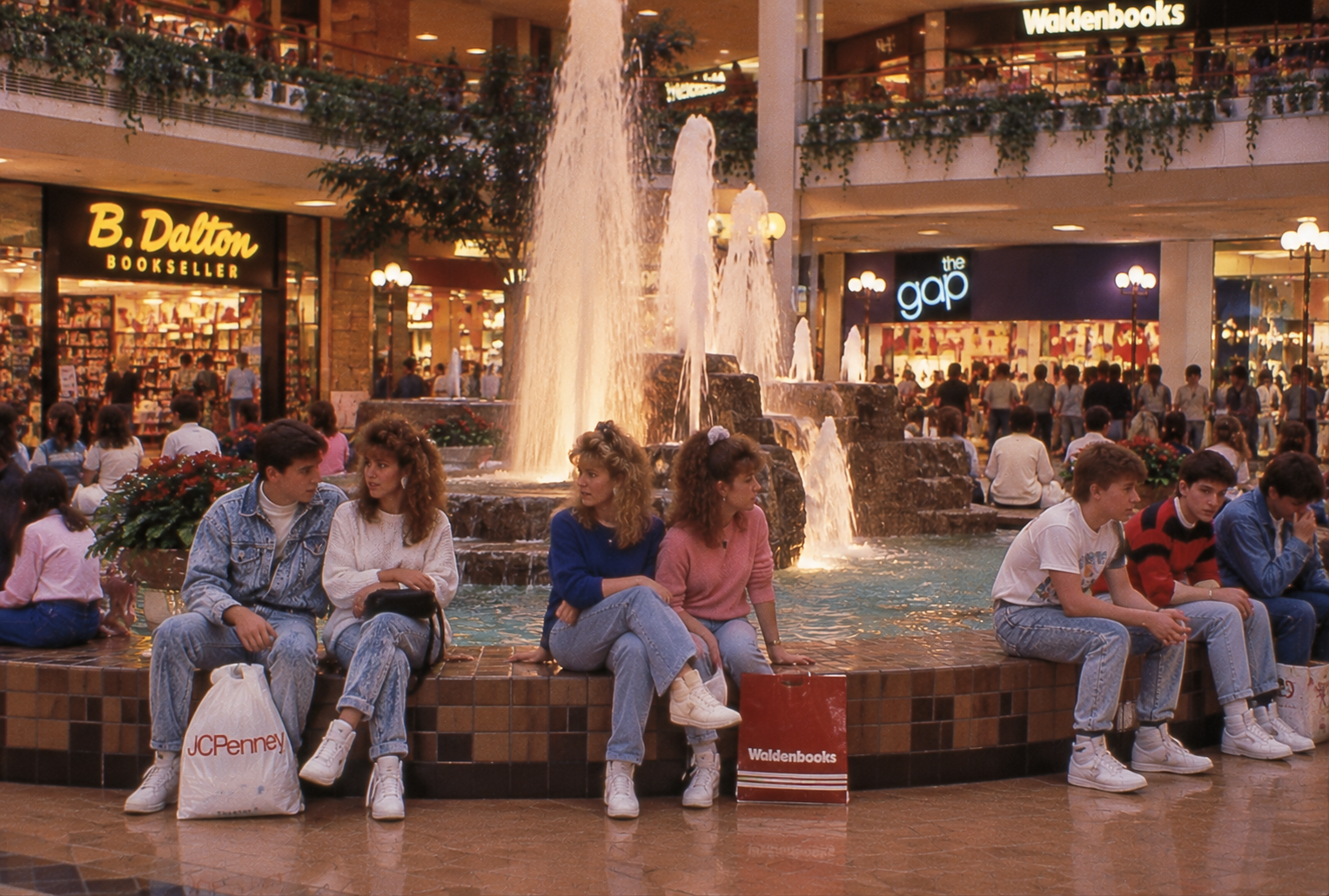 Teens in 1980s fashion sit on the edge of a mall fountain near stores like B. Dalton, Waldenbooks, and The Gap, holding shopping bags and chatting. The mall is busy and brightly lit.