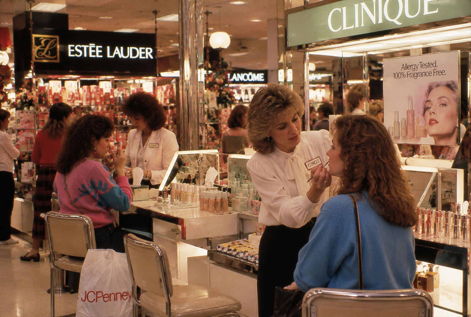 A Clinique beauty consultant applies makeup to a seated customer at a department store cosmetics counter. Estee Lauder and Lancome displays are visible, along with shoppers and a JCPenney bag in the scene.