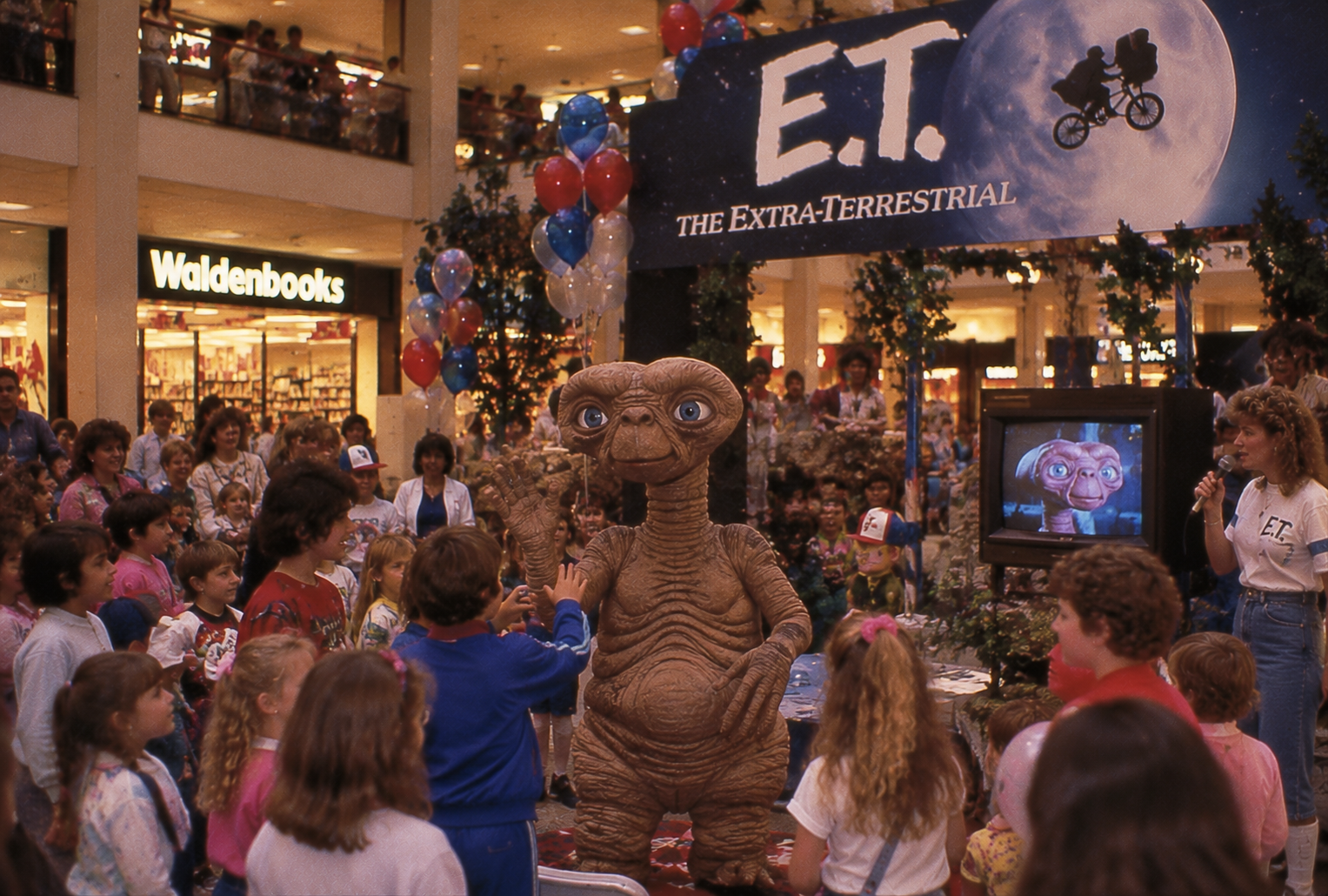 E.T. mascot greets children at a mall event promoting “E.T. The Extra-Terrestrial,” surrounded by balloons, decorations, and a crowd near a Waldenbooks store. A TV shows E.T.’s face.