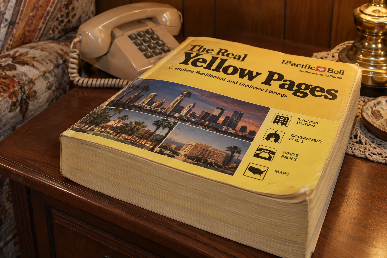 A large Pacific Bell Yellow Pages phone book sits on a wooden table next to a rotary phone and a lamp, showing a cityscape on the cover.