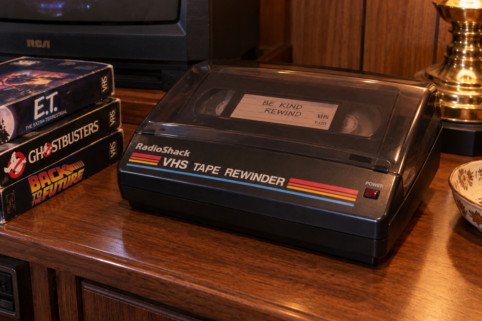 A VHS tape rewinder labeled “RadioShack” sits on a wooden table next to VHS tapes of E.T., Ghostbusters, and Back to the Future. The tape inside says, “Be Kind Rewind.” A lamp and TV are also visible.