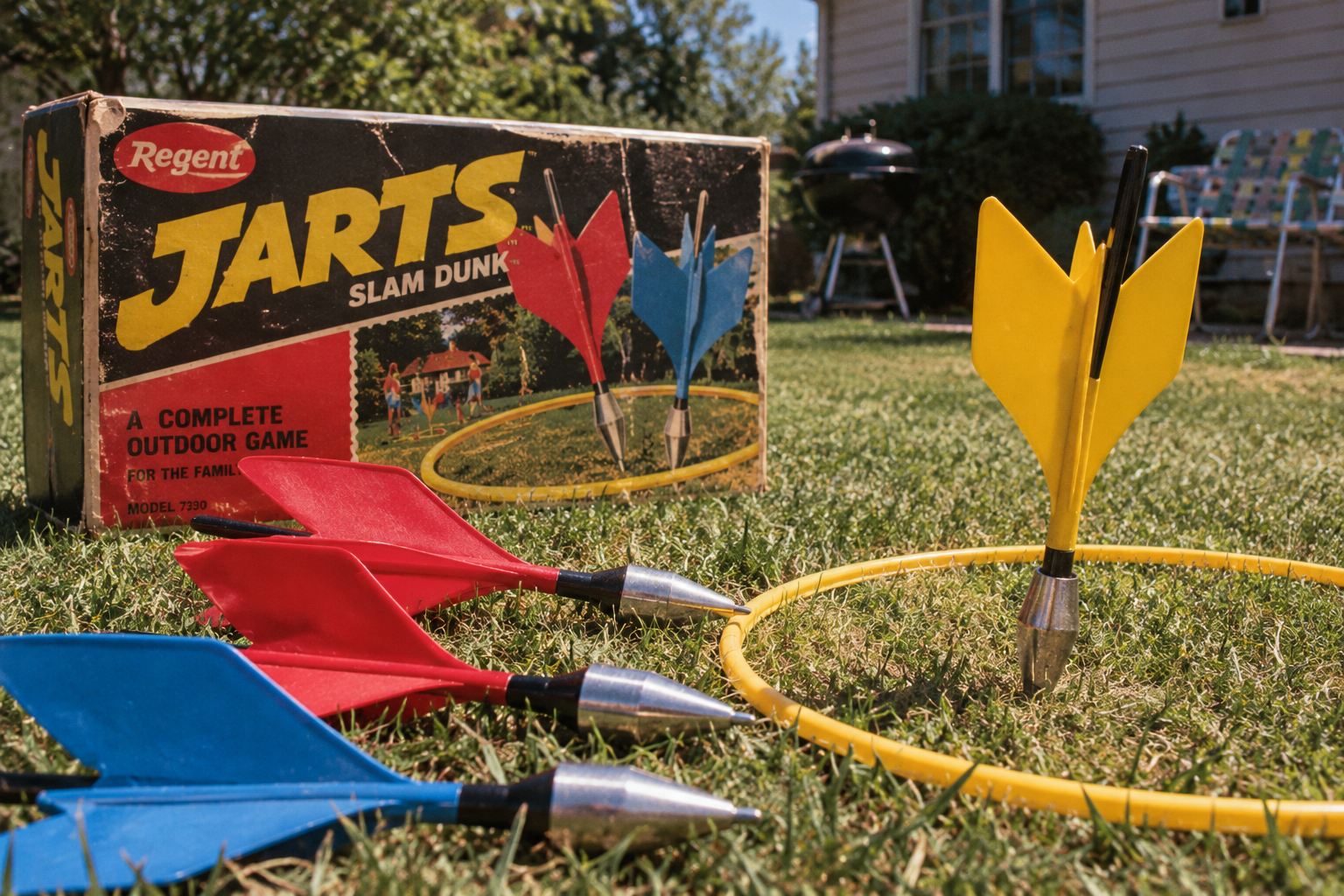 Four lawn darts, three red and one yellow, rest on grass near a yellow hoop. A Regent Jarts Slam Dunk game box is in the background along with a grill and house.