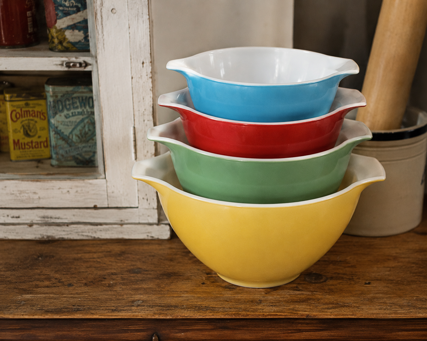 Four vintage mixing bowls in yellow, green, red, and blue are stacked on a wooden table beside a white cabinet with old tin containers.