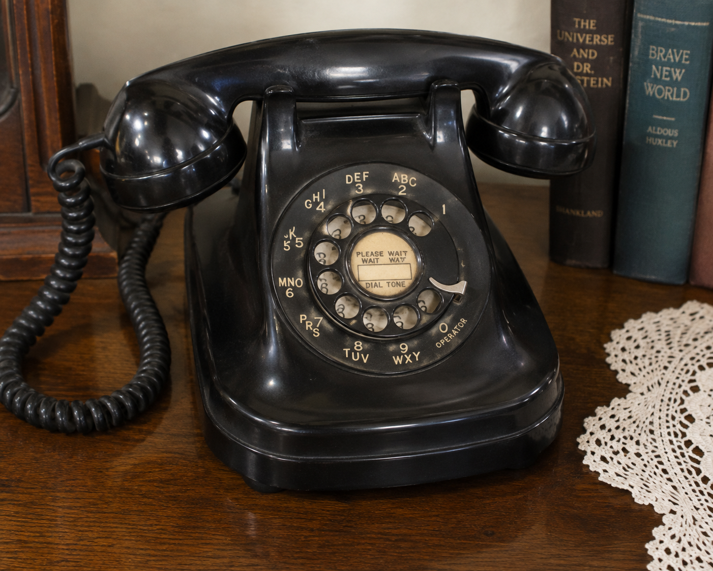 A vintage black rotary dial telephone sits on a wooden table next to books and a doily.