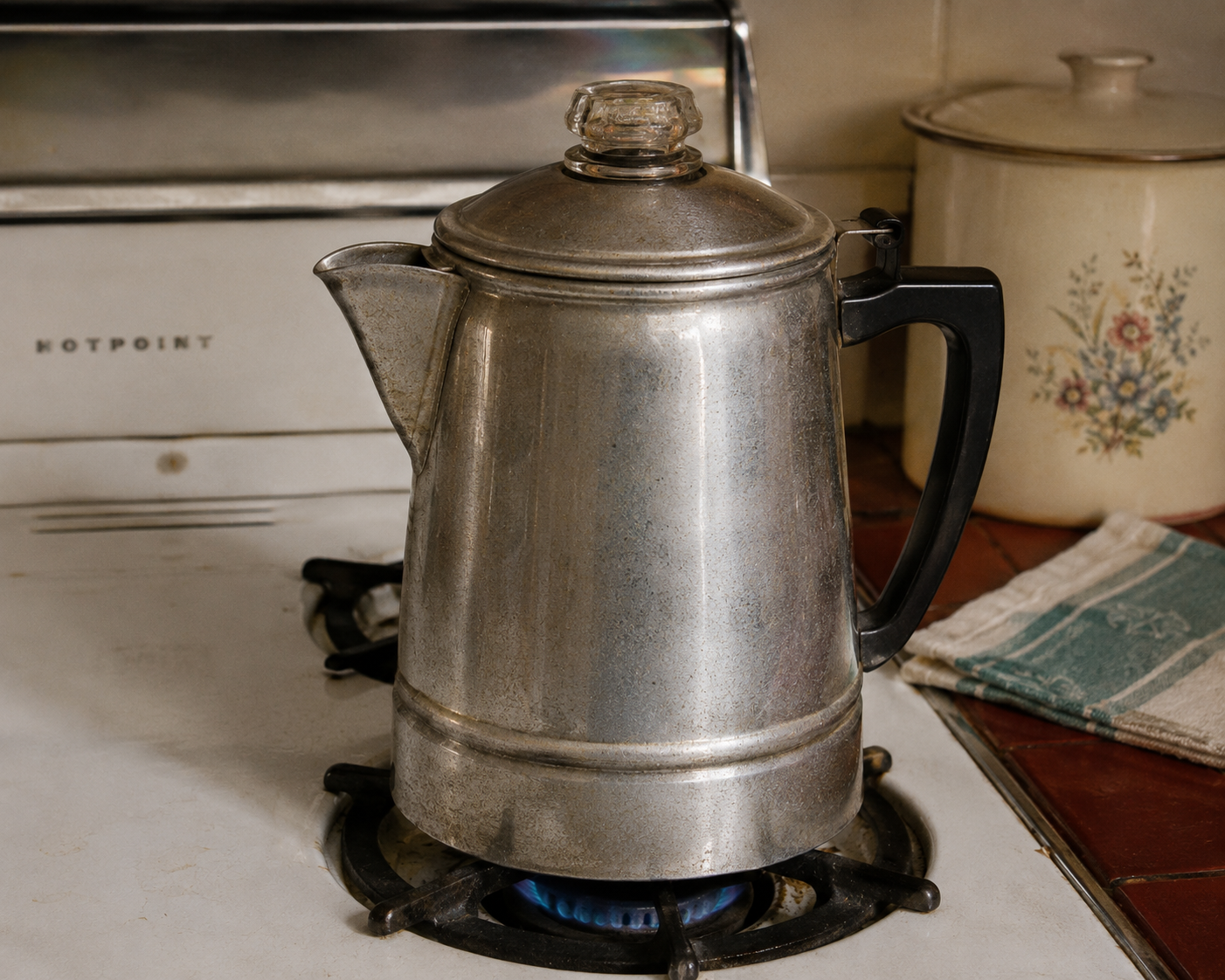 A vintage metal percolator sits on a gas stove burner with a blue flame. A beige container with a floral design and a green-striped towel are nearby on the countertop.