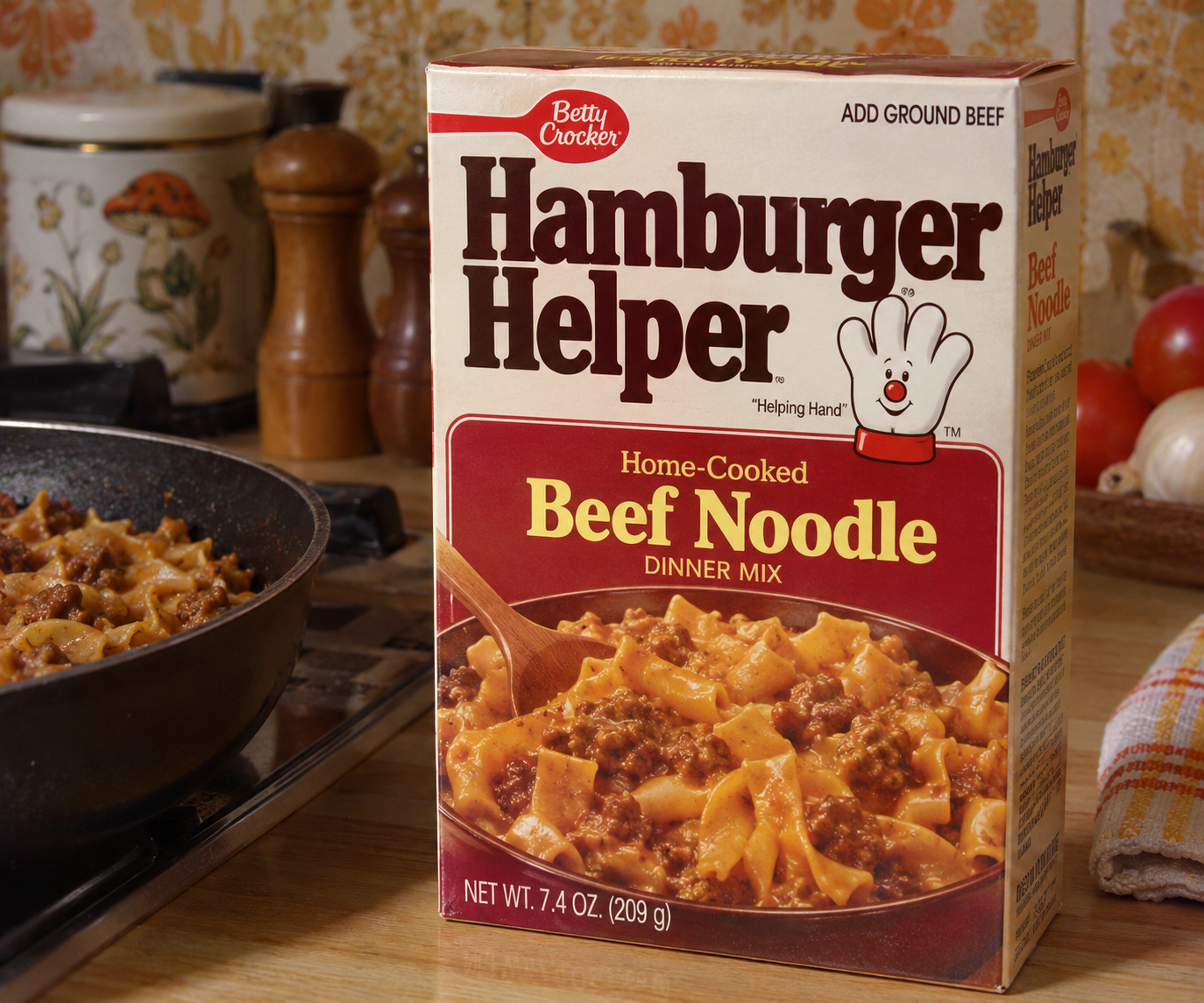 A box of Betty Crocker Hamburger Helper Beef Noodle dinner mix sits on a kitchen counter next to a pan of prepared food. The kitchen background includes jars, a towel, and onions.