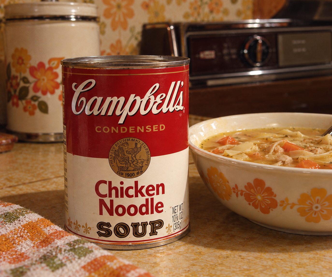 A can of Campbell’s Chicken Noodle Soup sits next to a bowl of soup on a retro kitchen counter with floral decor in the background.