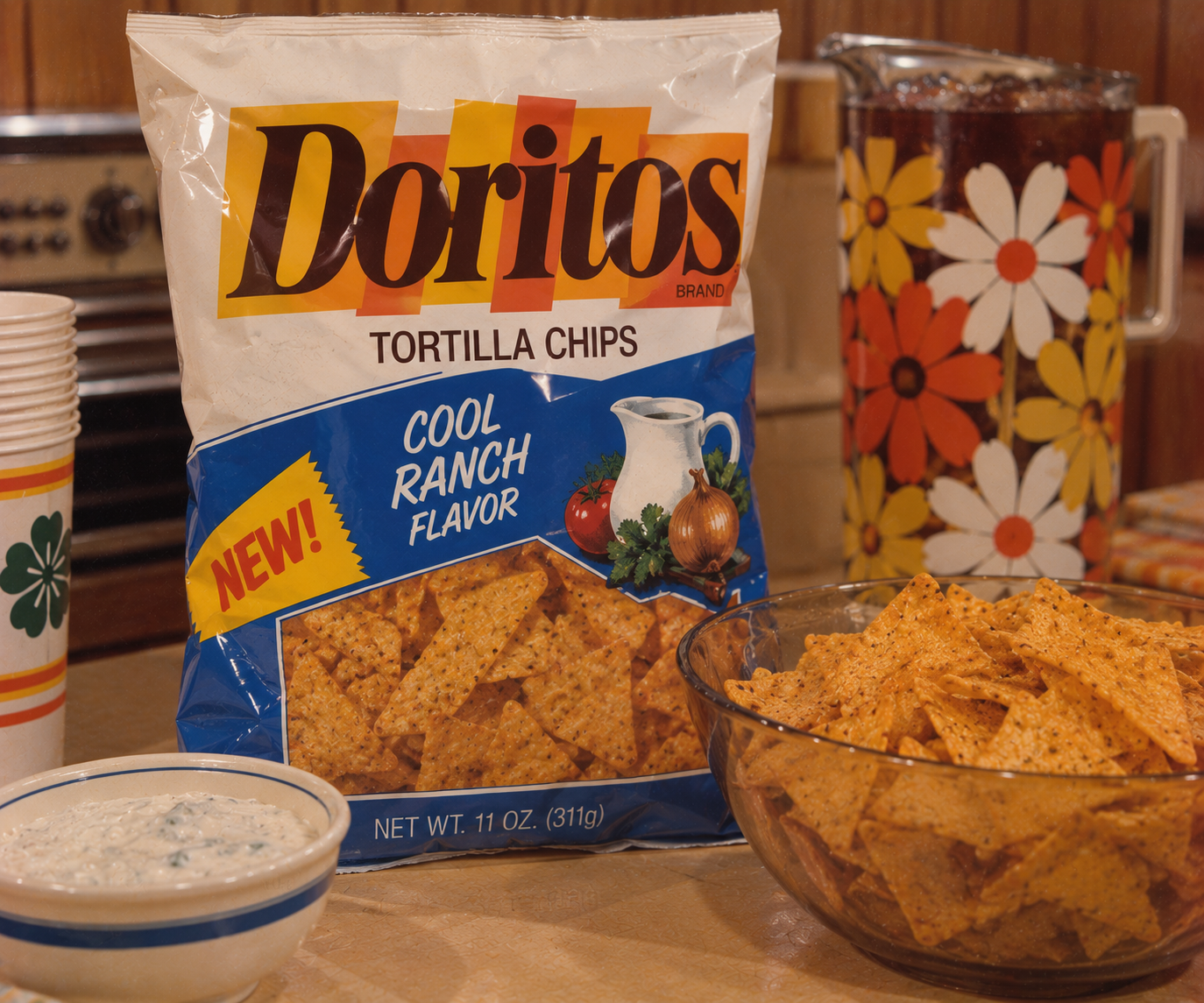 A bag of Cool Ranch Doritos, a true 1980s grocery store staple, sits on a table next to chips, dip, stacked cups, and a floral pitcher in a retro kitchen setting.