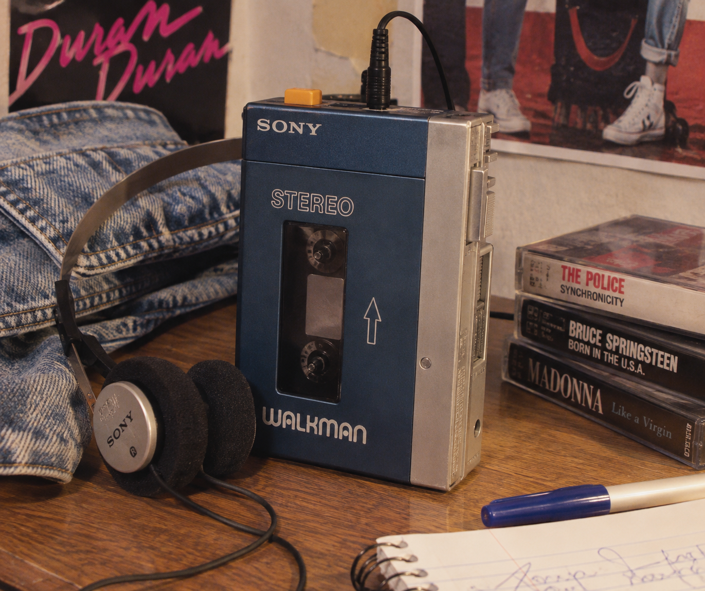 A blue Sony Walkman with headphones sits on a desk next to cassette tapes, a denim jacket, and posters of 1980s music artists on the wall in the background.